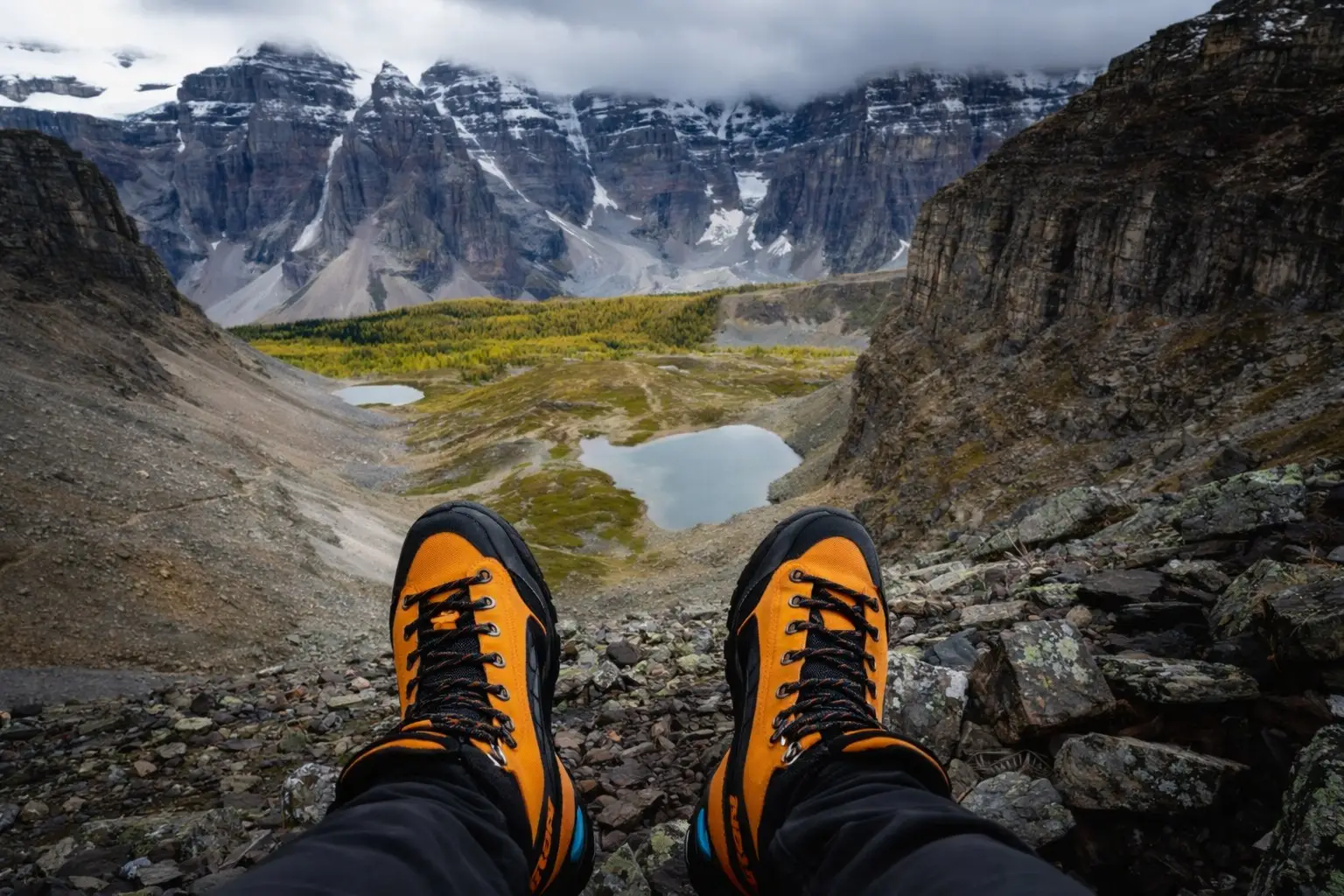 A first-person, eye-level perspective taken from the top of Sentinel Pass in Banff National Park. The hiker's orange and black mountain boots are in the immediate foreground, overlooking the sprawling Larch Valley below, which is filled with vibrant yellow larch trees and small alpine ponds, set against a backdrop of jagged, snow-capped mountain peaks under a cloudy sky.