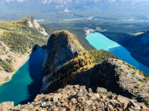 A dramatic, high-altitude panoramic view from the summit of the Devil's Thumb in Banff National Park. In the foreground, a rugged, rocky ridge descends toward the valley floor. To the left is the deep, dark blue Lake Agnes, and to the right, the brilliantly turquoise Lake Louise. The vast, mountainous landscape of the Canadian Rockies extends into the background under a clear sky.