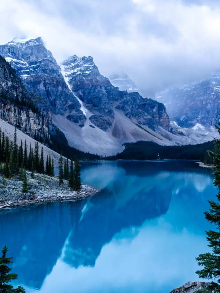 A stunning early morning landscape of Moraine Lake in Banff National Park, Alberta, taken from the famous Rockpile vantage point. The deep, still turquoise water perfectly reflects the towering, snow-dusted jagged peaks of the Valley of the Ten Peaks. Dense evergreen forests line the shore, and a soft, misty morning light covers the rugged mountain faces.