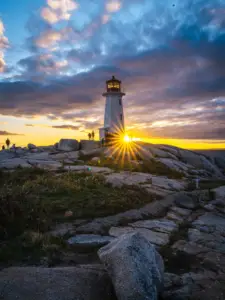 A landscape photograph of Peggy's Cove Lighthouse in Nova Scotia at sunset, with a golden sunstar flare behind the lighthouse and a cloudy sky.