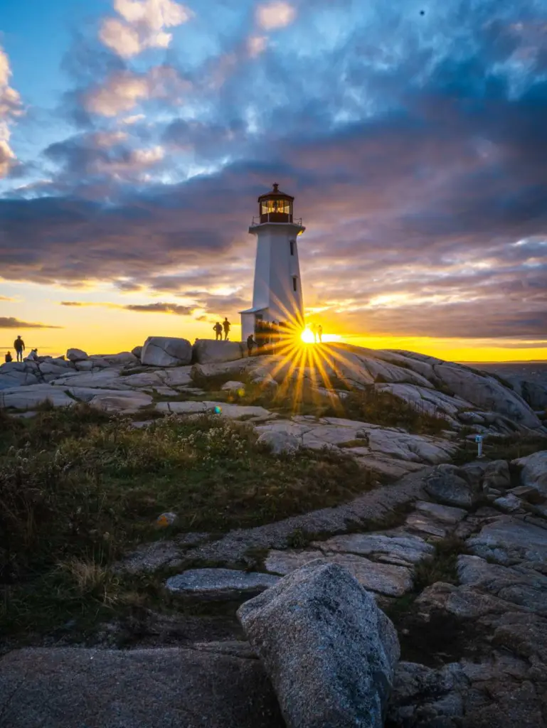 A landscape photograph of Peggy's Cove Lighthouse in Nova Scotia at sunset, with a golden sunstar flare behind the lighthouse and a cloudy sky.