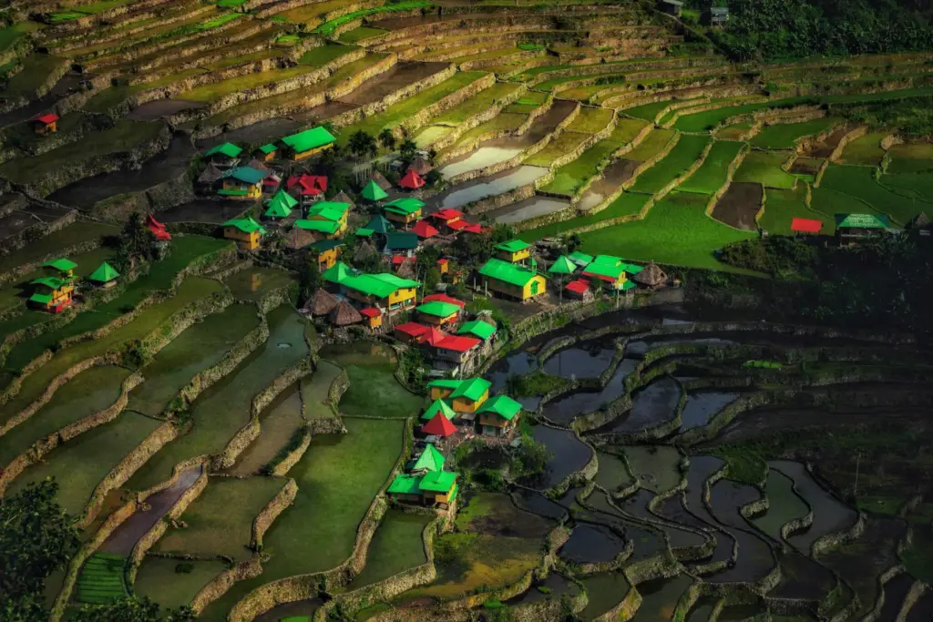 A bird's-eye view of the Batad Rice Terraces in Ifugao, Philippines. The photo captures the intricate, carved patterns of the lush green rice paddies forming a massive amphitheater shape, with a small cluster of traditional village houses with bright red and green roofs nestled in the center.