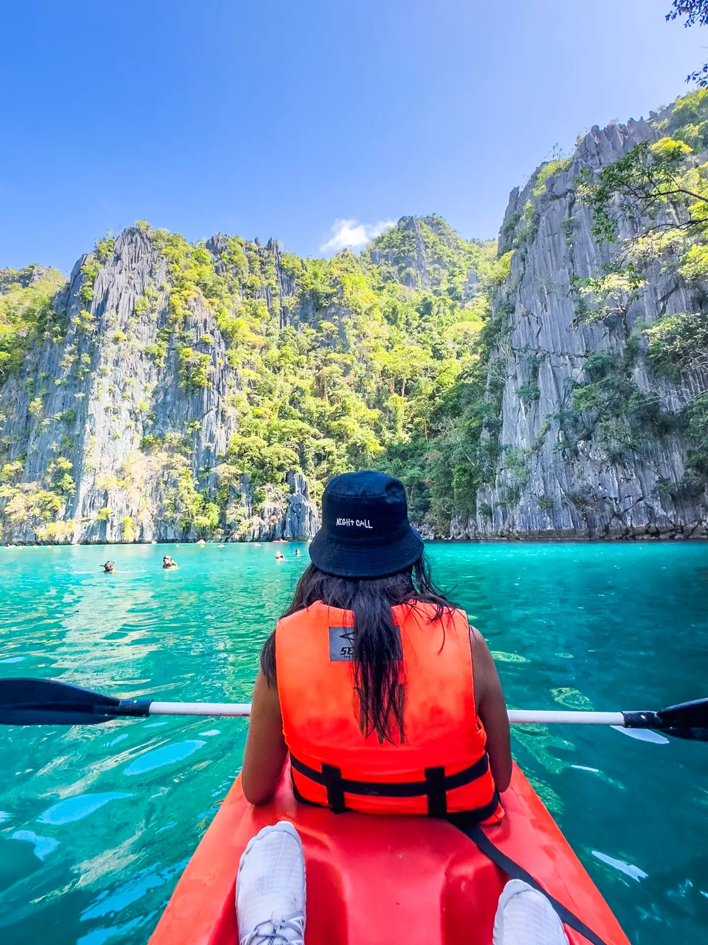 A first-person perspective from a red kayak inside the inner Twin Lagoon in Coron, Palawan. A person in an orange life vest and black bucket hat looks toward the towering, lush limestone karst cliffs and the vibrant turquoise water under a clear blue sky.