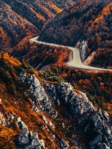 Aerial view from the Skyline Trail boardwalk looking east over the winding Cabot Trail highway. Rugged cliffs with orange and red autumn foliage drop steeply into the valley as cars drive the scenic road.