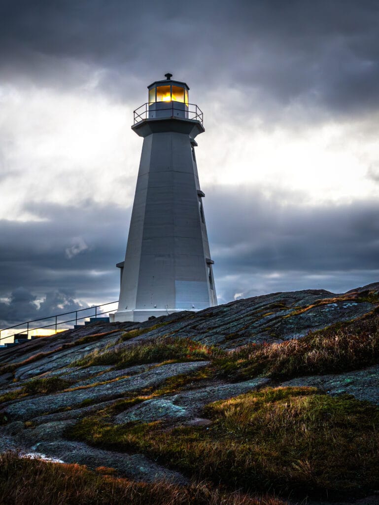 A tall, white lighthouse standing atop a rugged, rocky cliff at Cape Spear. The lantern at the top is glowing with a warm yellow light against a dark, dramatic sky filled with thick grey clouds. The foreground shows weathered, lichen-covered rocks and patches of coastal grass.
