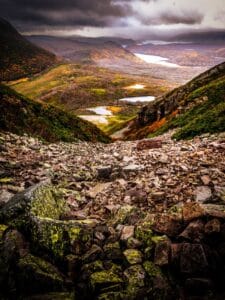 A steep, rocky ascent on the Gros Morne mountain trail, looking down into a valley with multiple alpine ponds. The scene is captured under a dramatic, dark, and cloudy sky with low-hanging fog, indicating an approaching storm.