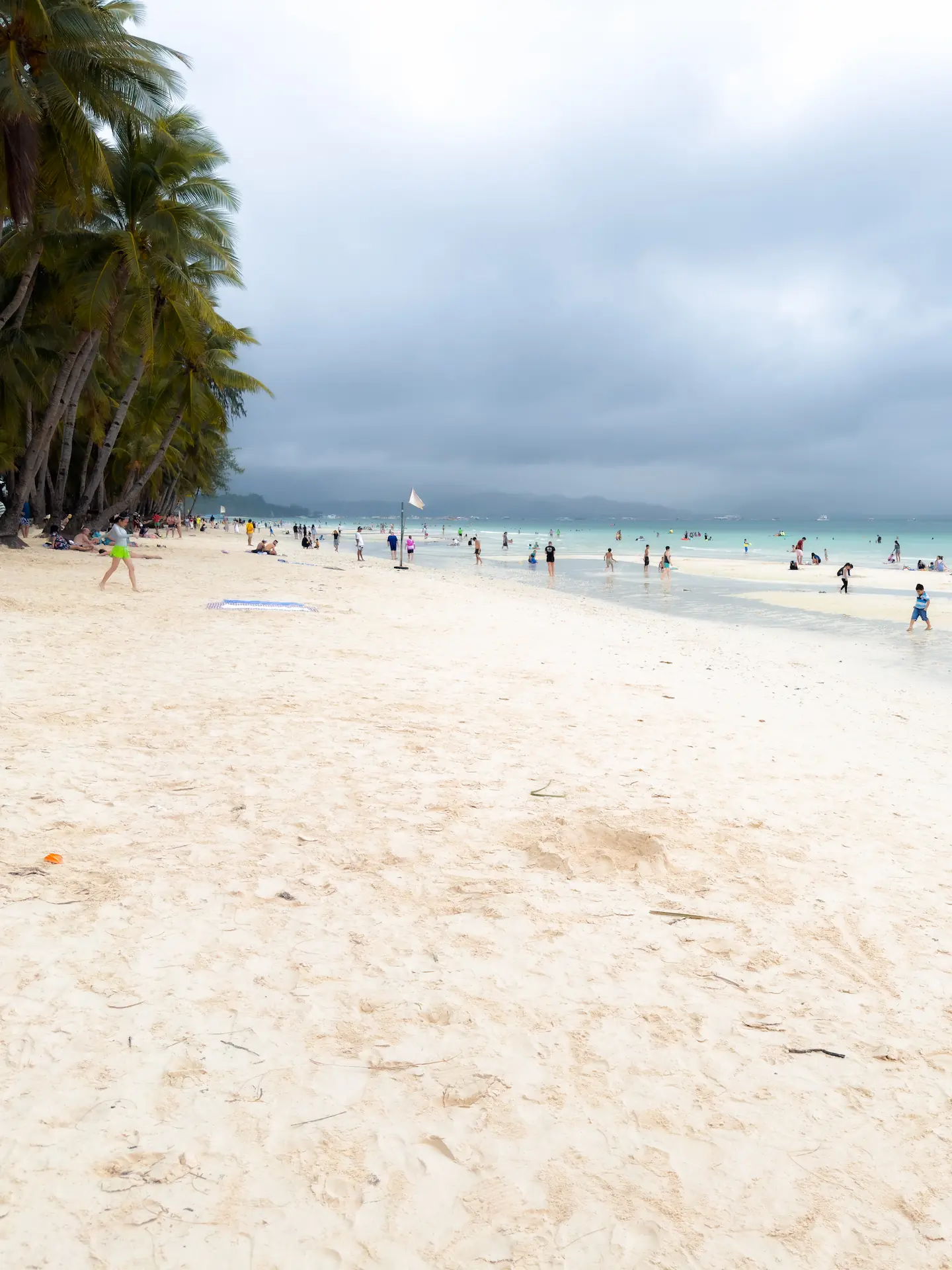 A scenic daytime view of White Beach in Boracay, Philippines, at Station 2. The beach is filled with soft white sand, people walking along the shoreline, and the turquoise water is visible under a cloudy sky, with dense palm trees lining the beach.