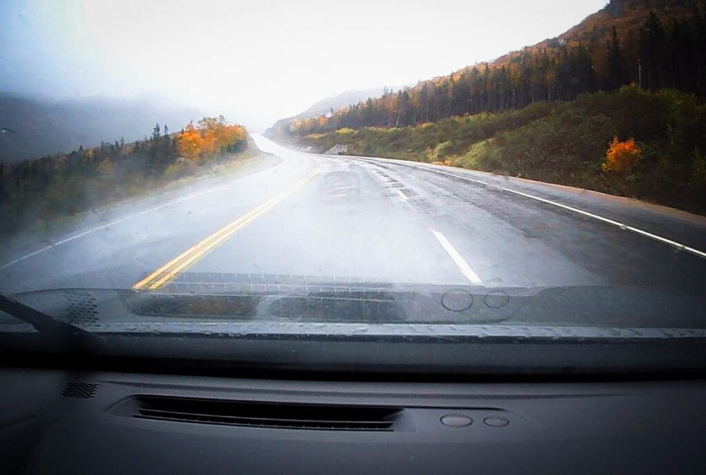 A first-person view from inside a car driving on a wet, winding highway toward Gros Morne National Park. The asphalt is slick with rain, reflecting the overcast sky, and the roadside is lined with autumn-colored trees and mist-covered hills.