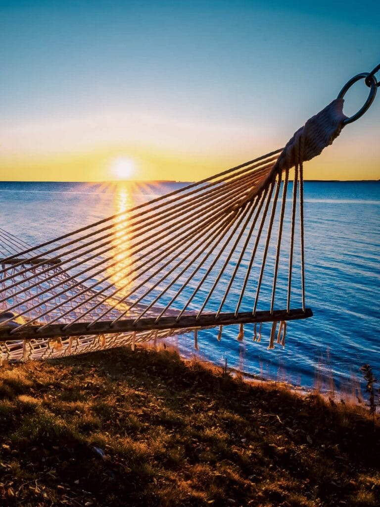A low-angle view of a white rope hammock on a grassy bluff at Sea Parrot Ocean View Manor. The morning sun rises over the calm Atlantic Ocean, casting a golden reflection on the water and lighting up the coast.