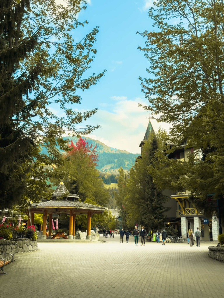Pedestrian Village Stroll in Whistler BC with wooden gazebo architecture and mountain peaks in the background under a blue sky.