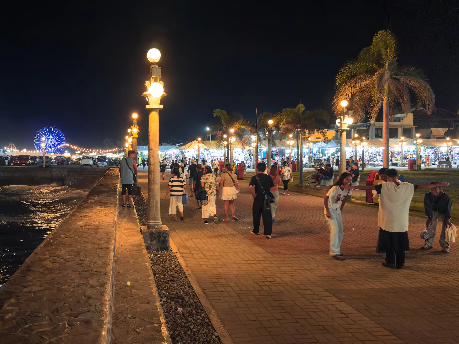 A vibrant night view of Rizal Boulevard in Dumaguete City featuring warm street lighting, people strolling, and a Ferris wheel in the background.