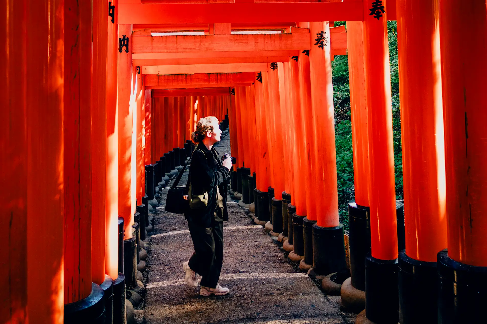 A traveler with a camera walks through the vibrant orange torii gates at Fushimi Inari Shrine during a Japan travel itinerary in Kyoto.