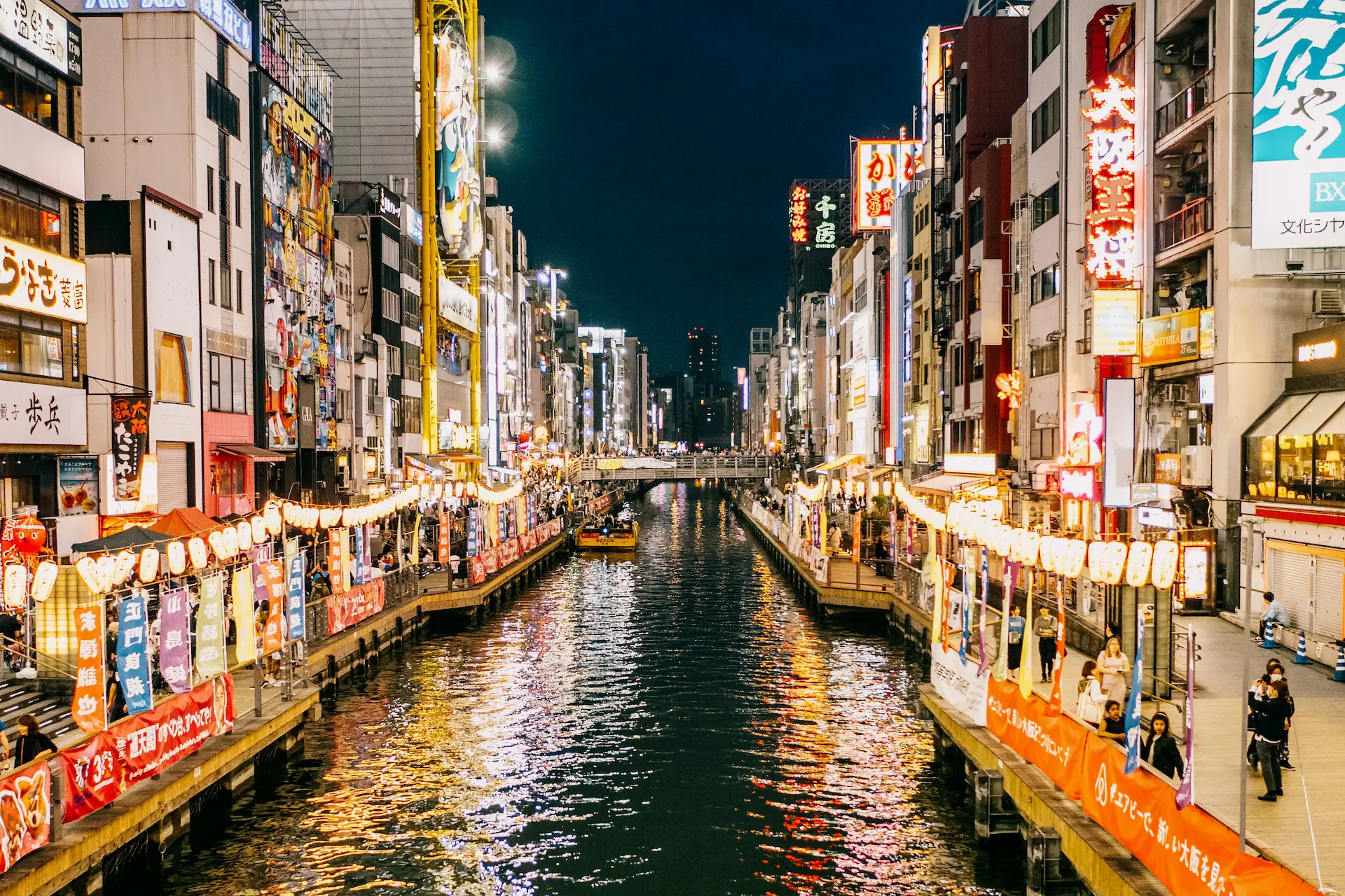 The brightly illuminated neon signs and colorful lanterns reflect on the Dotonbori Canal at night during a Japan travel itinerary in Osaka.